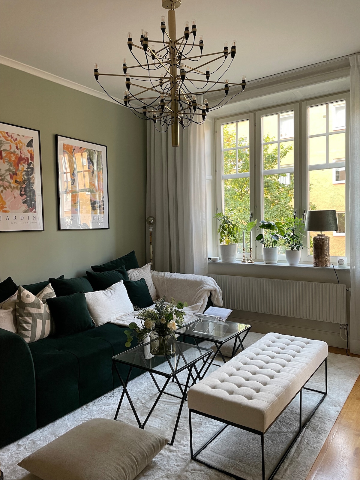 A welcoming living room features a dark green sofa adorned with decorative pillows. A glass coffee table sits in the center, accompanied by a plush bench. Large windows allow natural light to illuminate the space, framed by soft curtains and showcasing decorative plants on the sill.
