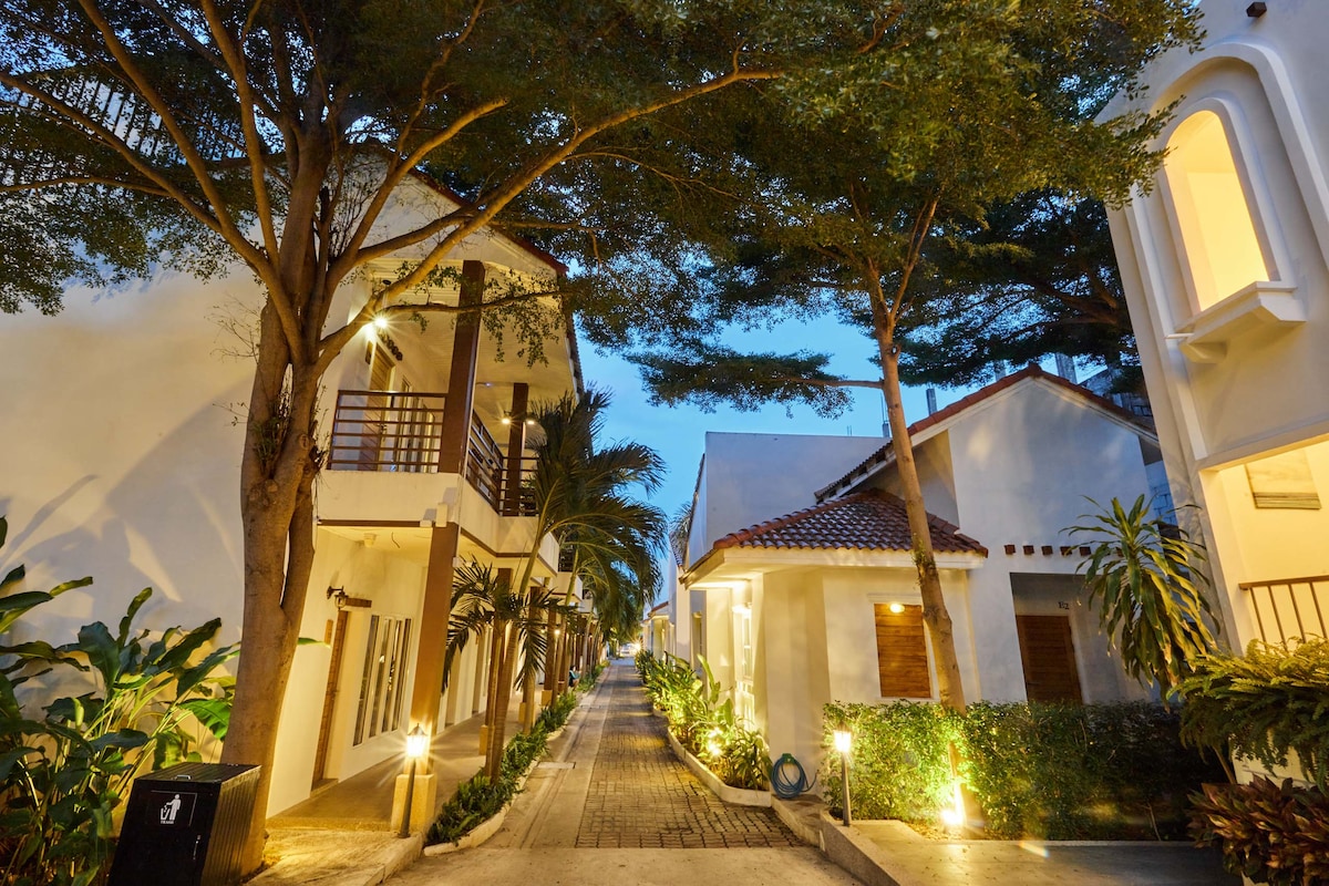 A serene walkway is flanked by two-story villas, surrounded by lush greenery and well-maintained gardens. Soft lighting highlights the path, creating an inviting atmosphere. The architecture features a blend of natural elements and contemporary design, contributing to a tranquil setting.