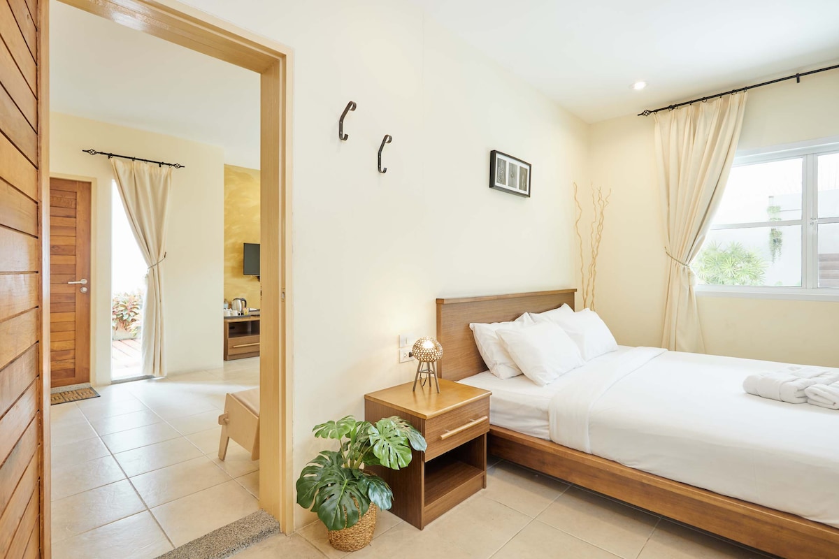 The bedroom features a spacious bed with crisp white linens and a bedside table. A decorative plant adds a touch of greenery, while natural light enters through a nearby window, complemented by soft curtains. A door leads to a connecting living area.