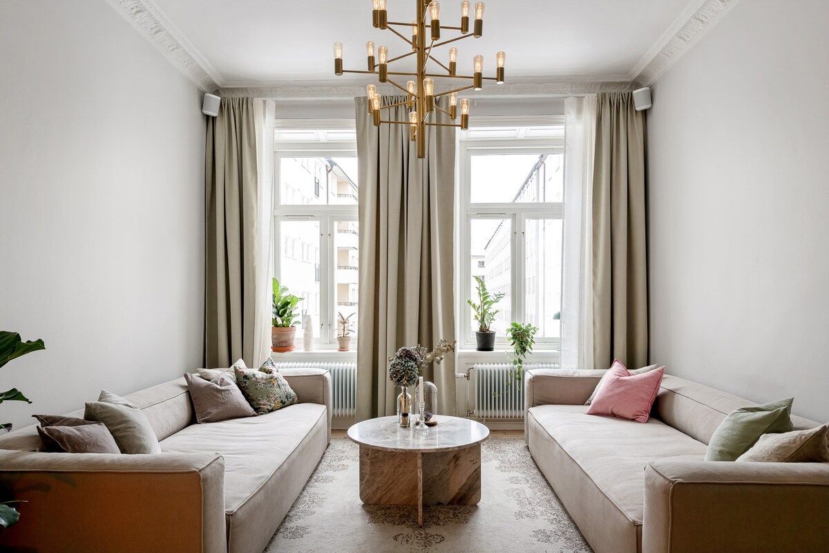 A serene living area features two spacious sofas adorned with decorative pillows. A marble coffee table sits at the center, complemented by natural light pouring in through large windows. Potted plants are positioned near the windows, adding a touch of greenery to the room.