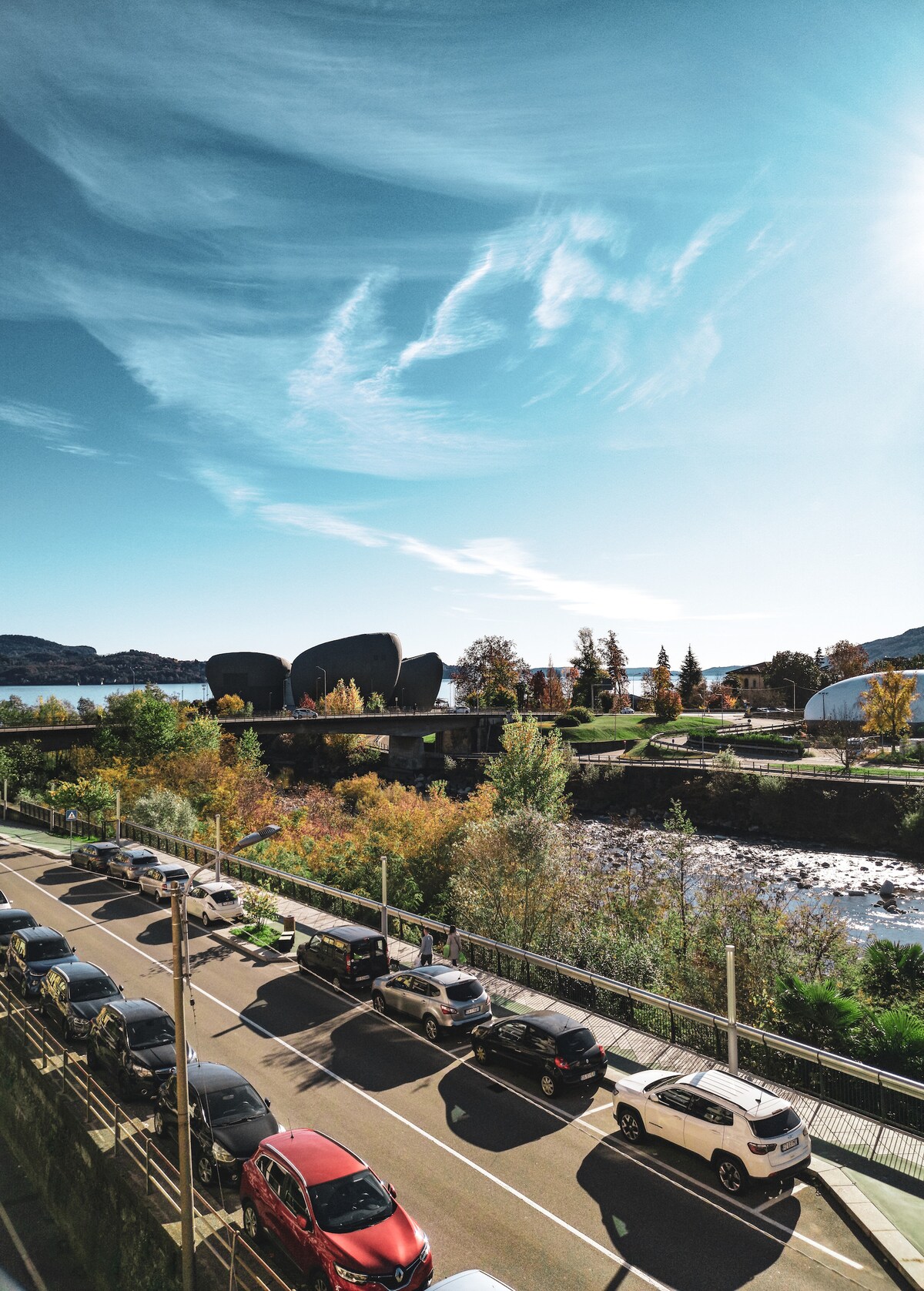 A view from the balcony showcases a serene landscape along the riverbank, with a picturesque mixture of trees in autumn colors lining a pedestrian path. Cars are parked along the street, with a modern building visible in the distance against a clear blue sky.