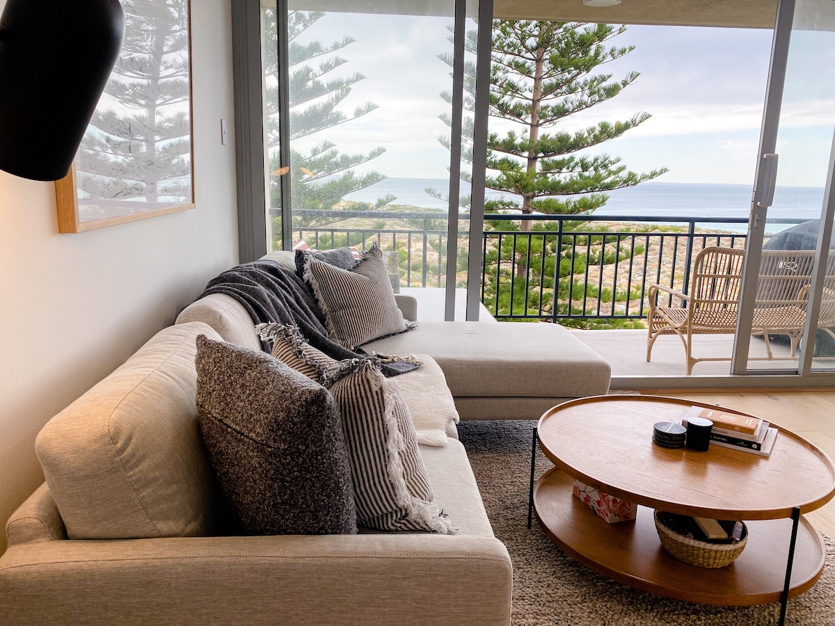 A spacious living area features a light-toned sectional sofa adorned with patterned cushions. A circular coffee table holds decorative items, while large glass doors open to a balcony with ocean views, framed by green trees in the background.