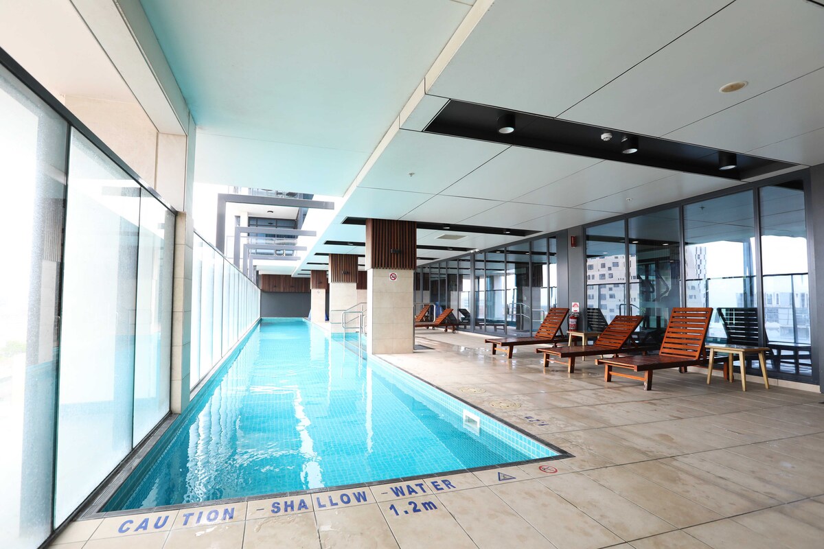 An indoor swimming pool is presented with a shallow water area indicated by signage. Sun loungers are arranged along one side, providing a space for relaxation. Glass walls allow for natural light to fill the space, enhancing the open atmosphere.