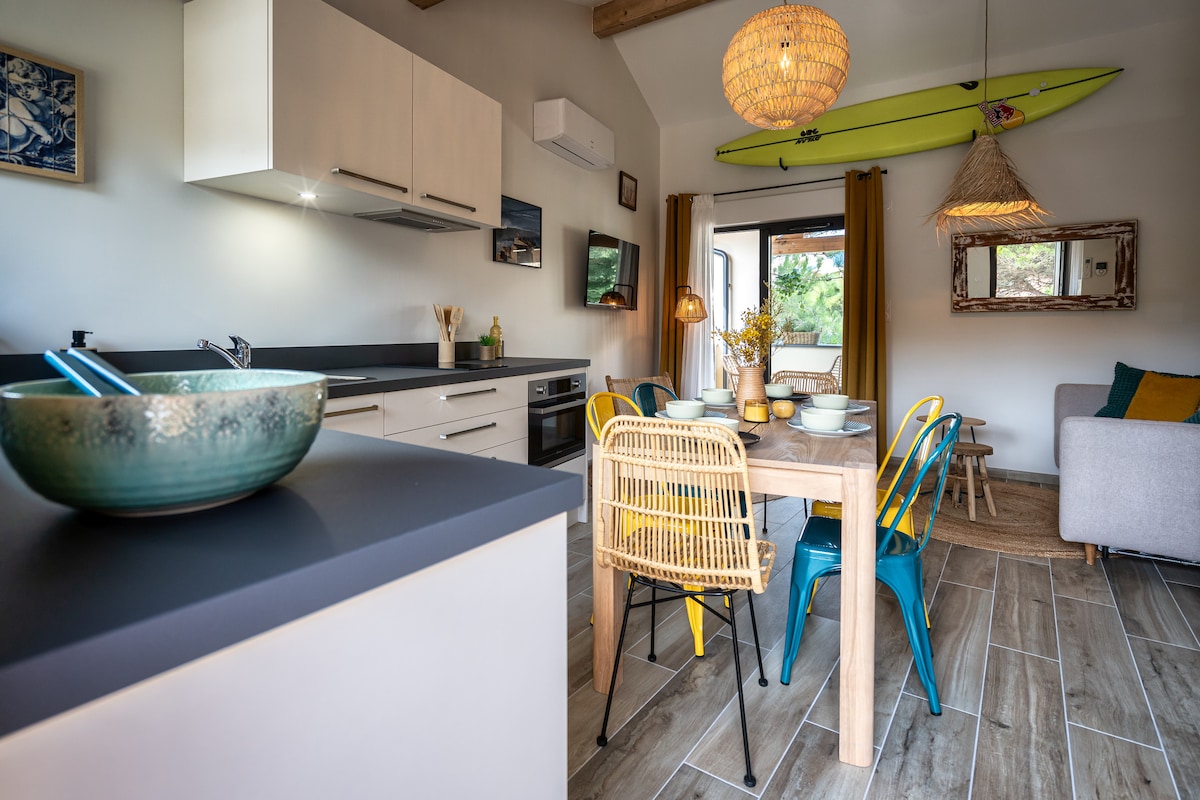 An open-concept kitchen and dining area features modern cabinetry and a light-colored countertop. A wooden dining table is surrounded by colorful chairs. A surfboard is mounted on the wall, while large windows allow for natural light and a view of the outdoor space.