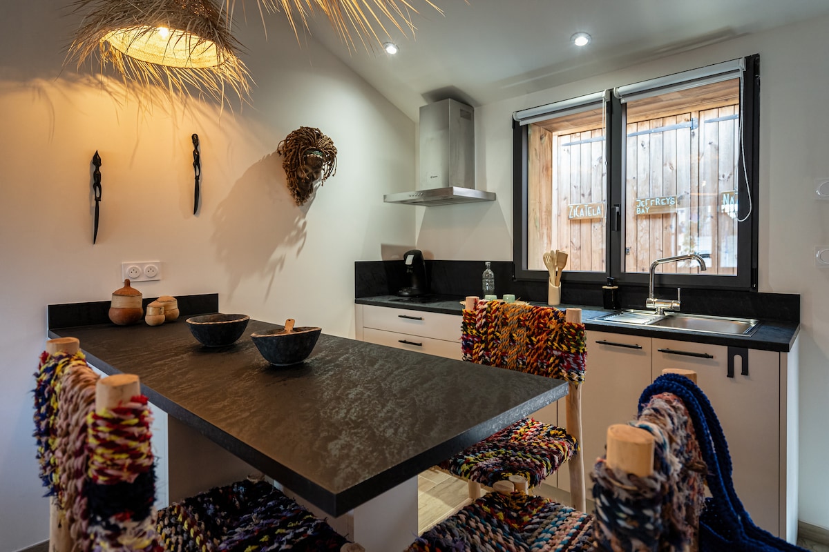 A modern kitchen is presented, featuring sleek black countertops and a spacious sink. Colorful woven chairs surround a dining table, bringing a vibrant touch to the space. Windows allow natural light to brighten the room while showcasing the external wooden elements.