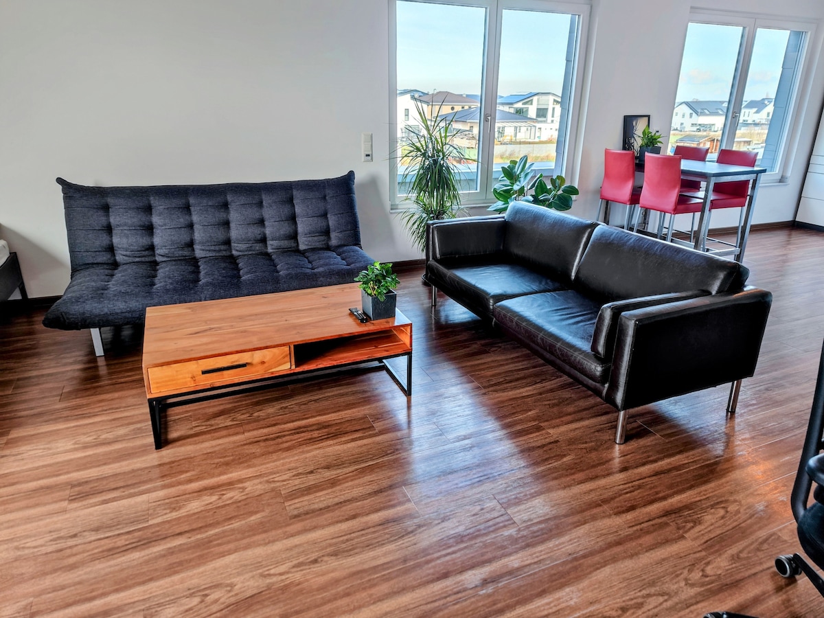 A modern living area is presented with a dark sofa, a black futon, and a wooden coffee table featuring a small potted plant. Large windows allow natural light to fill the space, complemented by a dining set in the background.