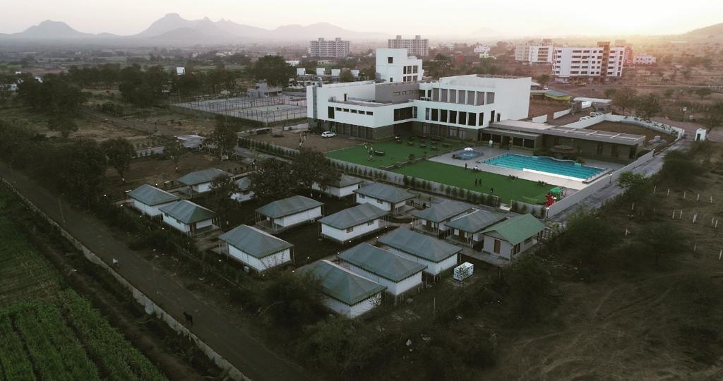 An aerial view captures the layout of the luxury tents and the main building. An olympic-sized pool is visible, surrounded by well-maintained lawns. In the background, mountains provide a scenic backdrop, while the surrounding landscape includes expansive fields and urban developments.