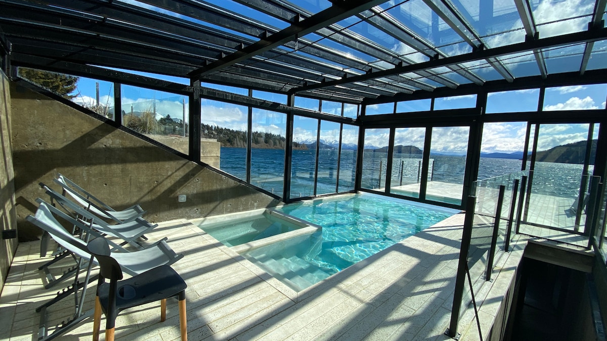 A modern indoor swimming area features a clear, heated pool and a jacuzzi, surrounded by expansive glass walls that provide stunning views of the lake. Sunlight streams in, illuminating the space, while sleek lounge chairs are positioned for relaxation.