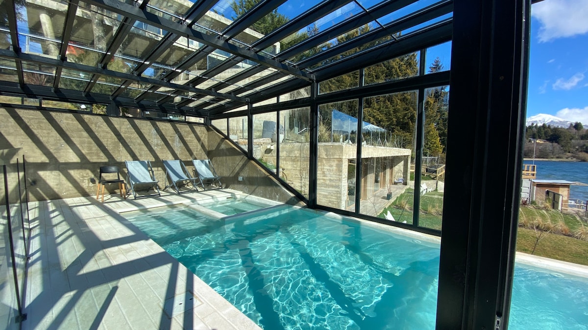 An indoor pool area is enclosed by expansive glass walls, allowing natural light to illuminate the space. Lounge chairs are positioned alongside the crystal-clear water, while the scenic outdoor views reflect the surrounding landscape and distant mountains.