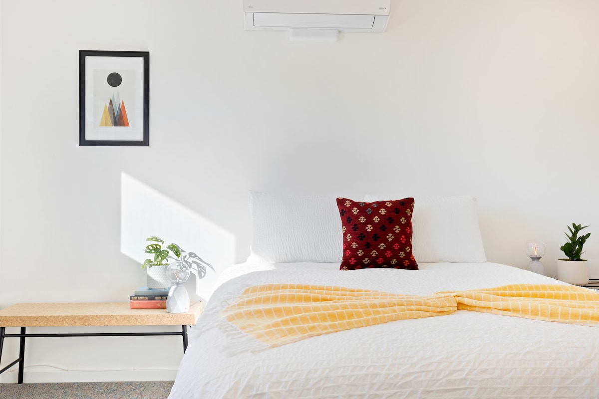 A bright and airy bedroom features a spacious bed with white linens and a decorative red pillow. A minimalistic artwork adorns the wall above the bed, while a small plant and candle are placed on a bench nearby, adding subtle accents to the calming space.