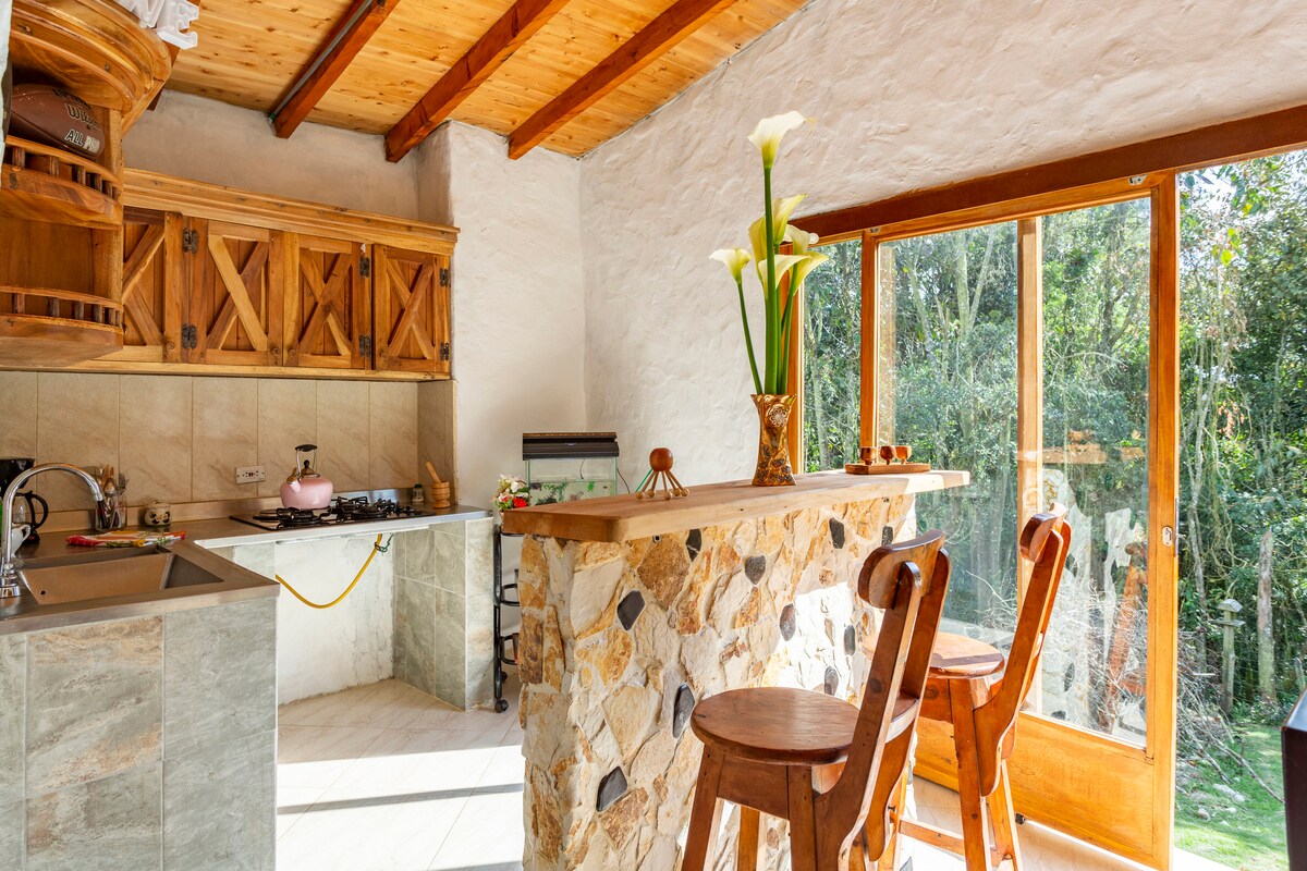 A rustic kitchen features wooden cabinetry and a stone-bar counter with two high stools. Large windows allow natural light to fill the space, with views of greenery outside. A gas stove is positioned on the countertop, alongside essential kitchen appliances.