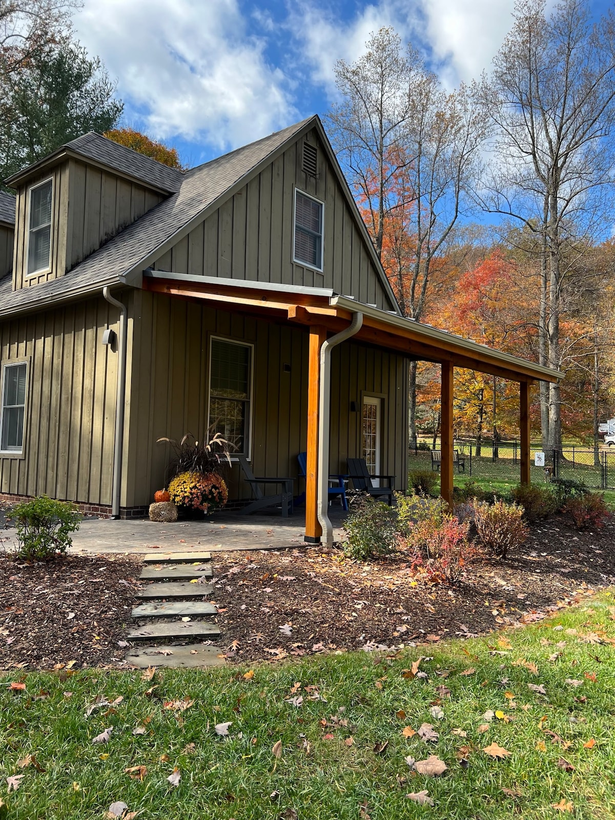 The exterior of a modern house is viewed, featuring a welcoming porch supported by wooden beams. Lush landscaping with shrubs and seasonal foliage surrounds the entry. The sky is partly cloudy, and trees with autumn leaves are visible in the background.
