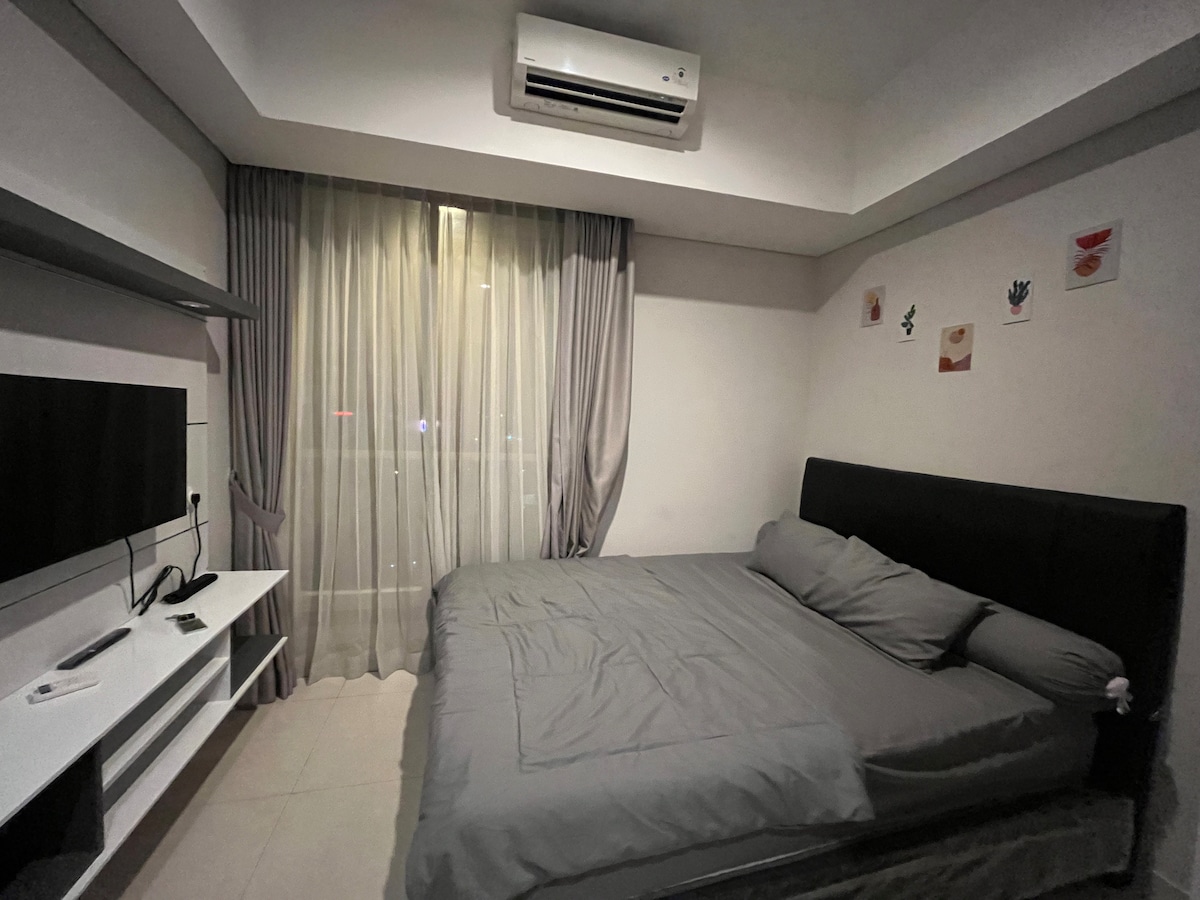 A cozy sleeping area features a spacious bed with gray bedding and a large window dressed with sheer curtains. A flat-screen television is mounted on the wall, and a minimalist shelf supports additional decor elements, providing a simple yet functional layout.