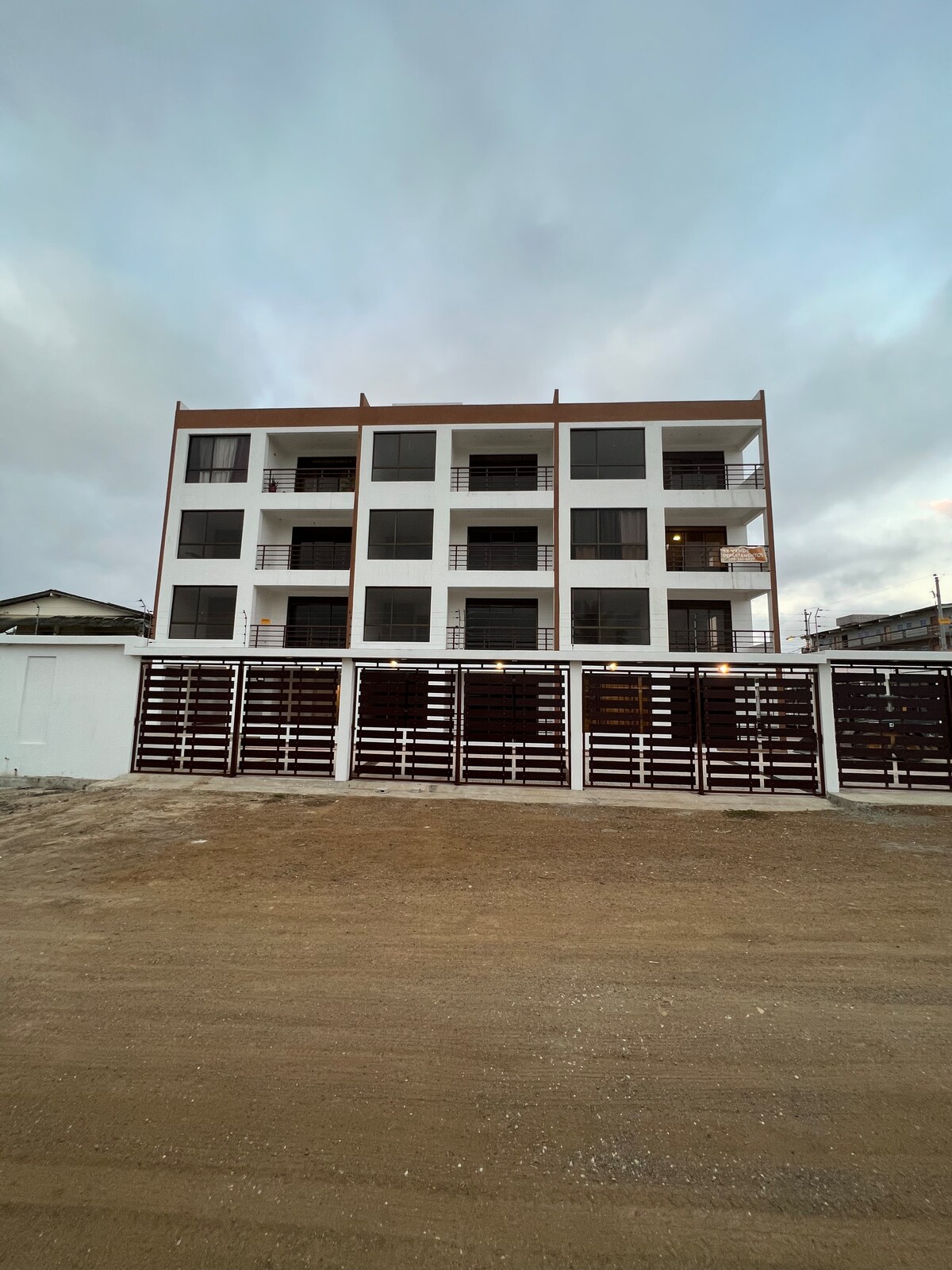 A modern, multilevel building features a clean design with large, rectangular windows. The structure is set against a cloudy sky, while a wooden fence with horizontal slats encloses the front. The surrounding area is mostly bare, with sand underfoot.