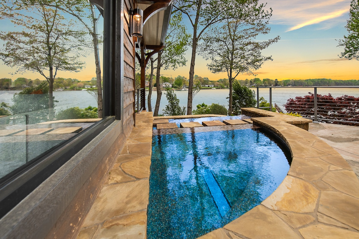 The image captures a serene pool area with a unique curved stone edge, overlooking a tranquil lake. The water reflects the sky's colors, while lush trees frame the view. A wooden railing and patio area are visible, enhancing the natural setting.