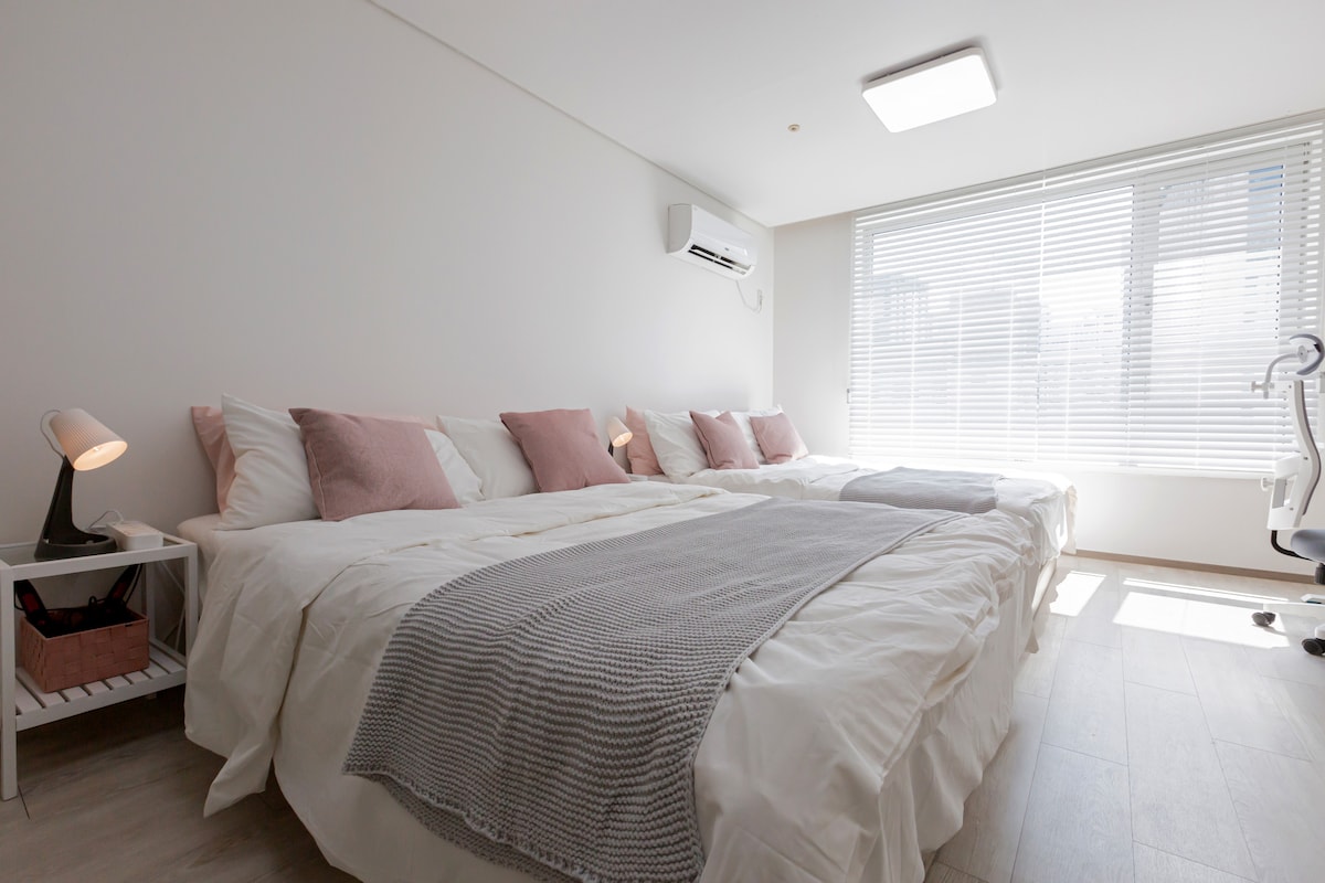 A well-lit sleeping area features two spacious queen size beds adorned with soft white linens and decorative pillows. A light grey blanket drapes over the foot of one bed. Natural light enters through a large window, complemented by adjustable blinds. A small bedside lamp and a desk chair are included.