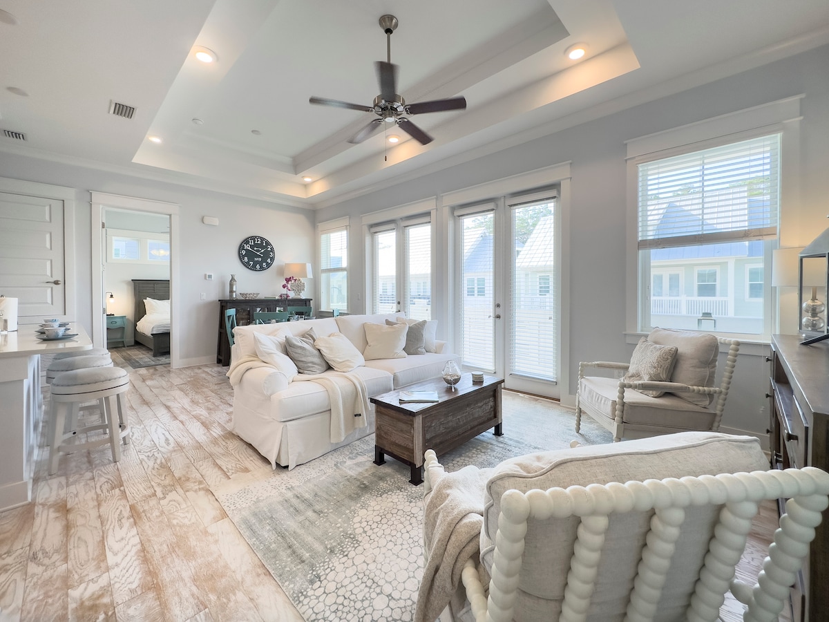 A bright and airy living area showcases large windows allowing natural light to fill the space. Comfortable seating, including a plush sofa and a rocking chair, surrounds a wooden coffee table. Pale wood flooring and an area rug add warmth to the modern decor.