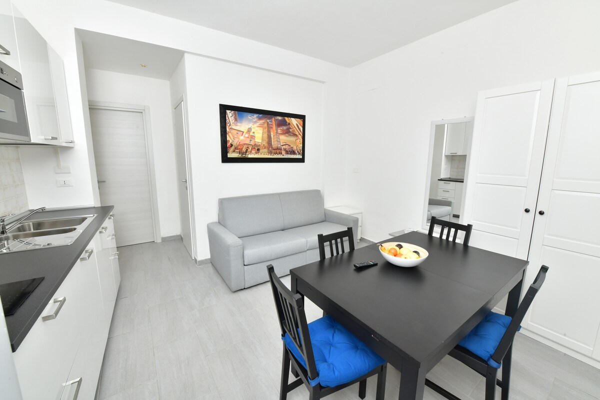 The open living space features a gray sofa positioned opposite a dining table with four black chairs. A bowl of fruit sits center stage on the table. The kitchen area, equipped with modern appliances, is visible in the background. Light-colored walls enhance the airy atmosphere.