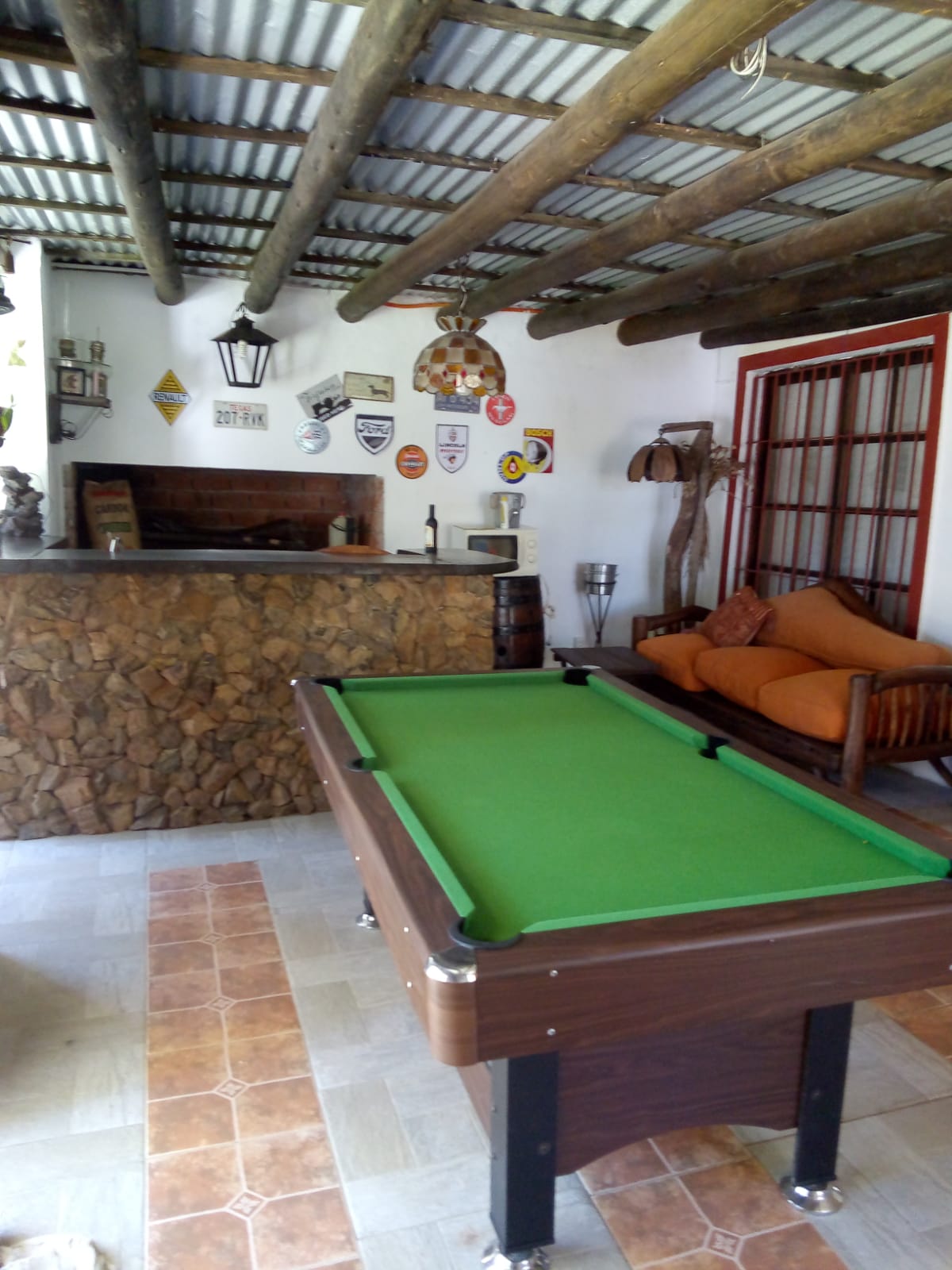 An entertainment area features a green felt pool table positioned centrally. A stone bar counter is visible in the background, adorned with various decorative items and memorabilia. Comfortable orange seating is arranged nearby, creating a relaxed space for social gatherings.