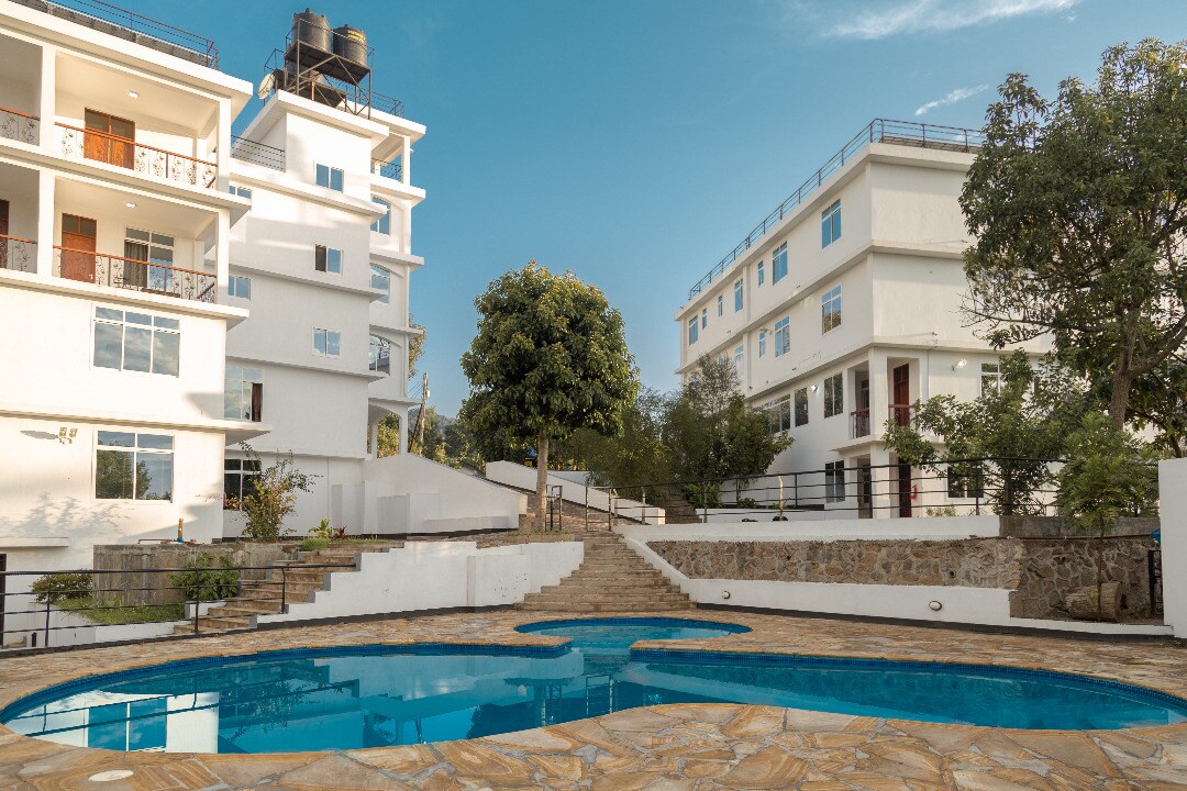 A shared outdoor pool is presented, bordered by natural stone and set against a backdrop of modern multi-story buildings. Lush greenery is visible around the pool area, enhancing the spaciousness of the surrounding environment. Clear blue skies add to the inviting atmosphere.