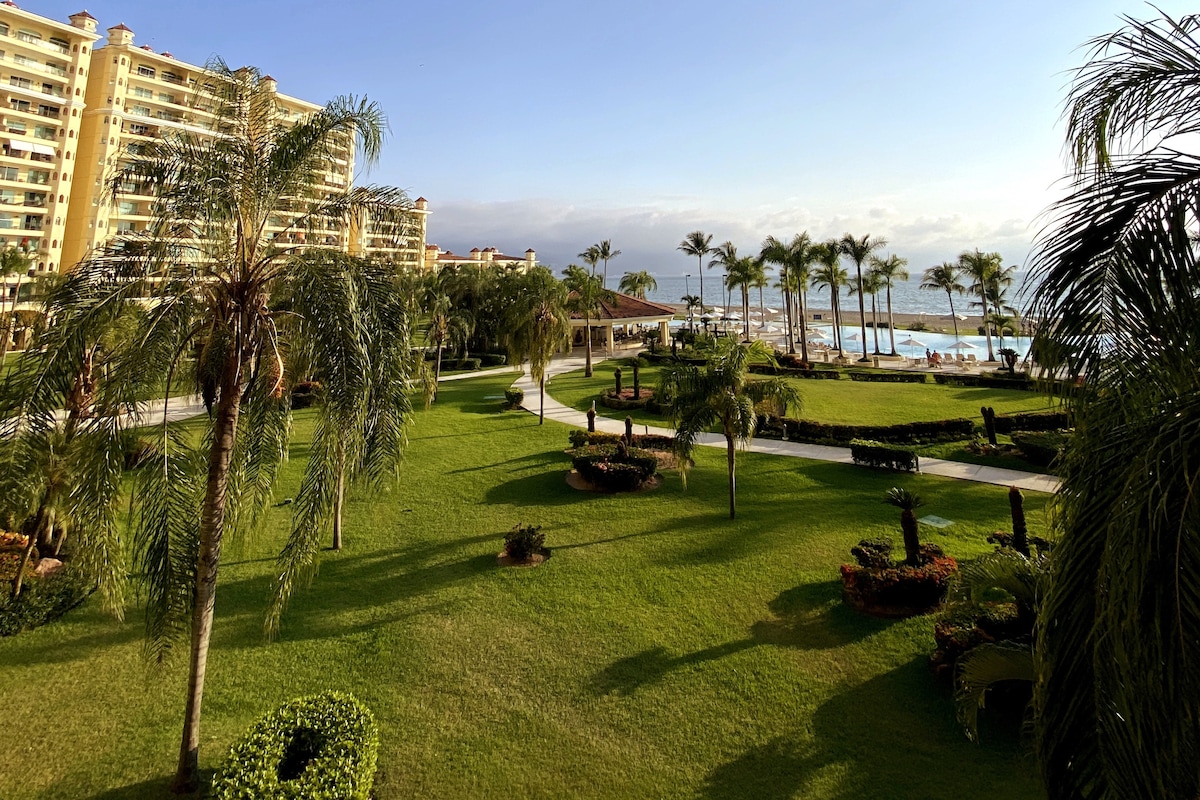 An expansive green lawn is framed by well-maintained gardens, featuring palm trees and neatly arranged shrubs. The view extends towards a sandy beach and a distant ocean, with a hotel structure visible along the horizon under a clear sky.