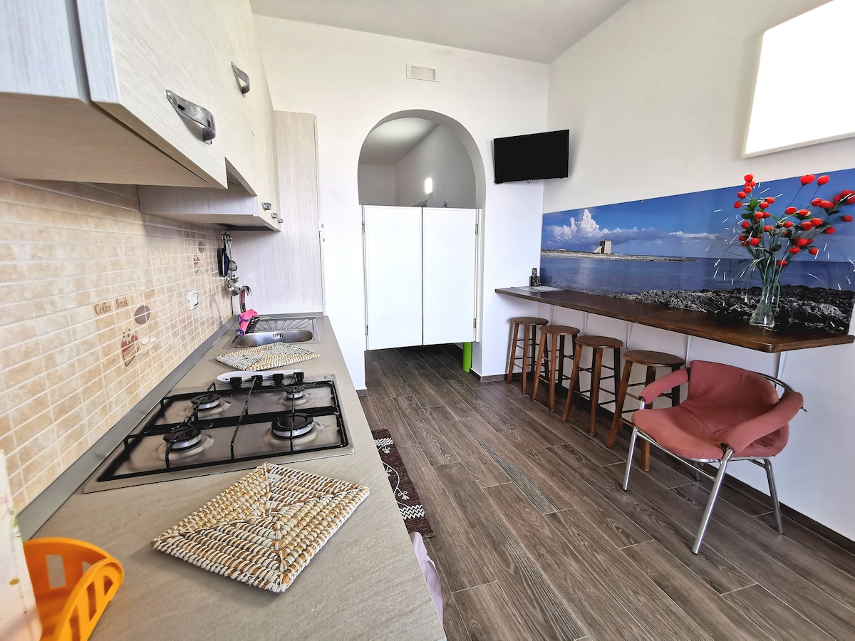 A functional kitchen features modern cabinetry and a gas stove. Barstools provide seating at a small table, while a wall-mounted TV is visible. A large photograph of a coastal scene decorates the wall, adding a touch of character to the space.