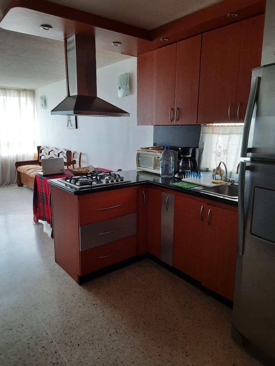 The kitchen features wooden cabinetry with a contemporary design. A stainless steel refrigerator and gas stove are positioned near a window that allows natural light. An adjacent seating area includes a small sofa and decorative items, creating a functional space.