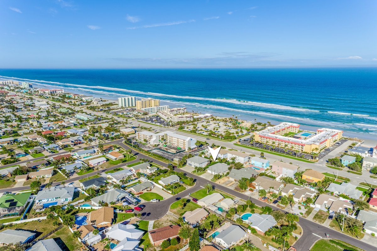 An aerial view showcases a coastal community, with sandy beaches stretching along the shoreline. Colorful buildings and homes are positioned among lush greenery. The vibrant blue ocean contrasts with the clear sky, emphasizing the inviting atmosphere of this vacation destination.
