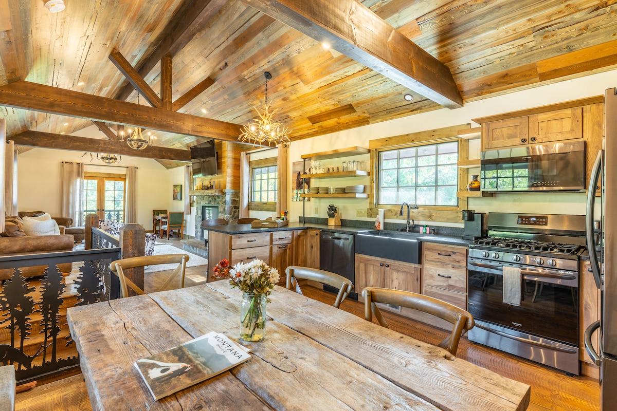 An open-plan living area features a rustic wooden dining table surrounded by light wooden chairs. The kitchen, equipped with modern appliances, displays dark countertops and storage shelves, while natural light pours in through large windows adorned with neutral curtains.