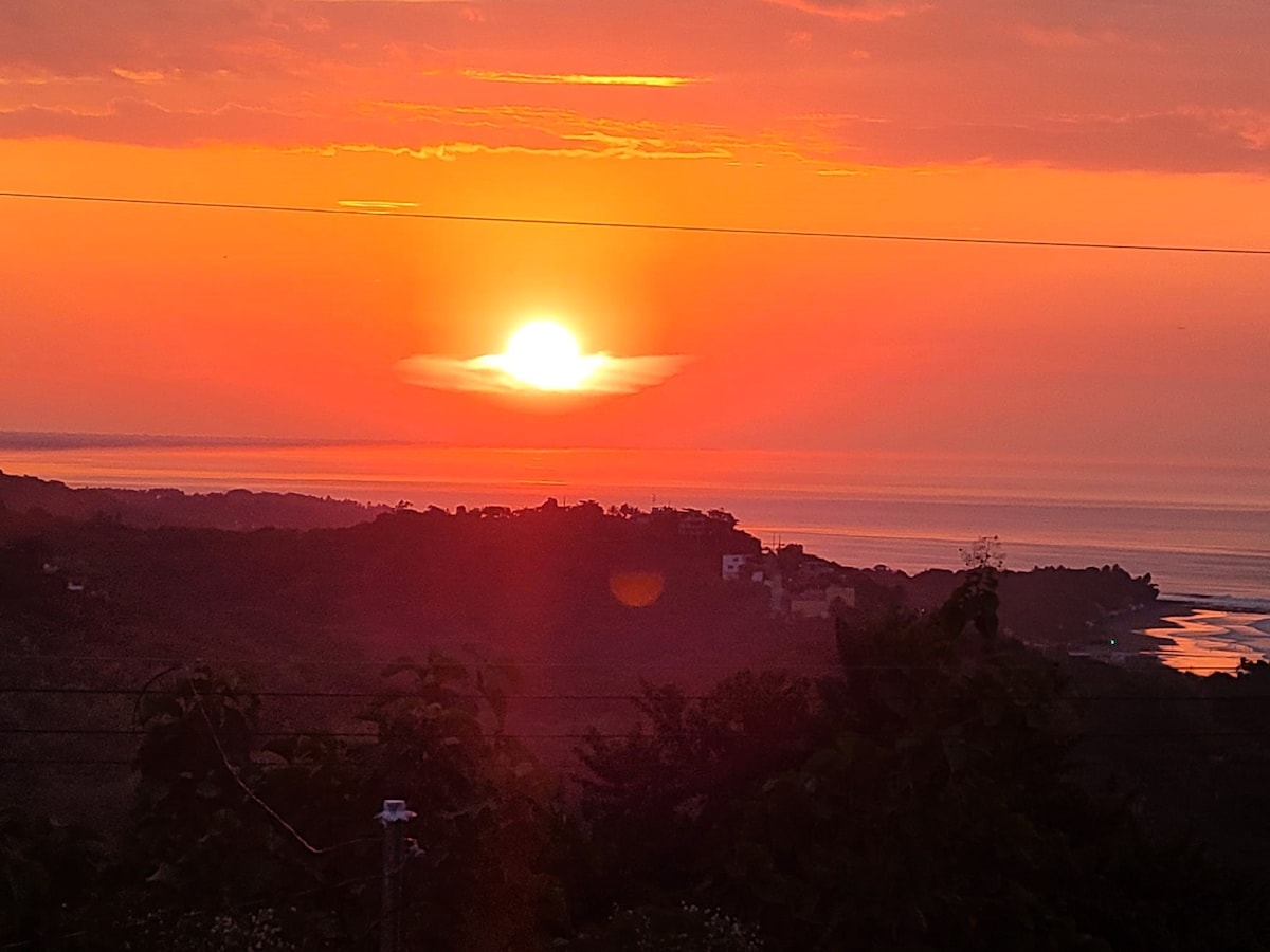 A vibrant sunset casts a warm orange glow over the ocean horizon, with silhouettes of distant hills framing the scene. The tranquil water reflects the colors of the sky, creating a serene atmosphere as the day transitions into evening.
