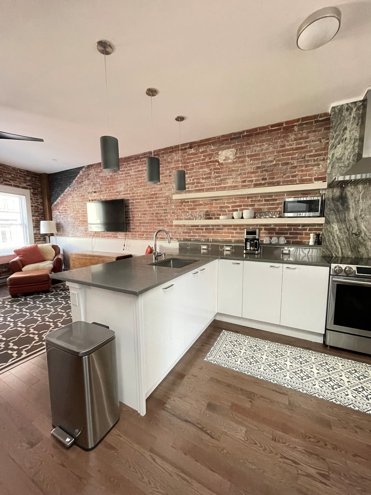 The kitchen space showcases a mix of modern and rustic elements, featuring stainless steel appliances, a sleek countertop, and a brick accent wall. Open shelving displays kitchen essentials, while a cozy seating area is visible in the background, highlighted by natural light from the window.