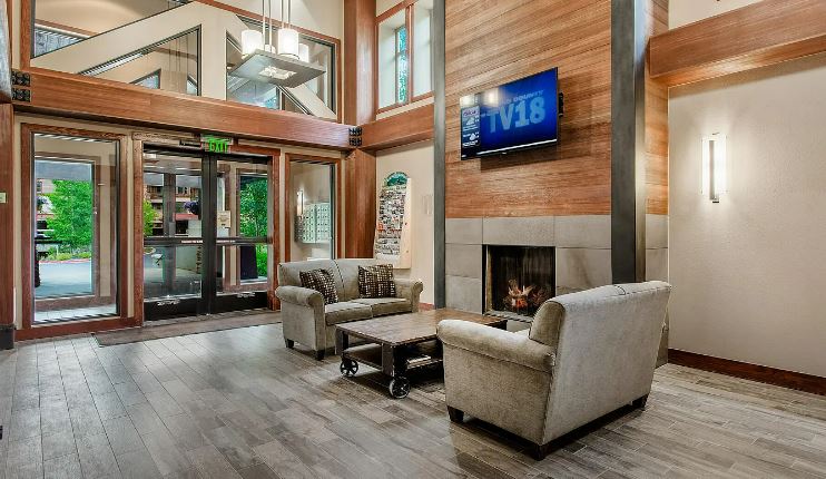 A spacious lobby area features two light-colored sofas arranged around a coffee table, with a modern fireplace set against a textured wall. Large windows allow natural light to fill the space, while a wall-mounted TV provides entertainment options. The flooring is a warm wood tone, enhancing the inviting atmosphere.