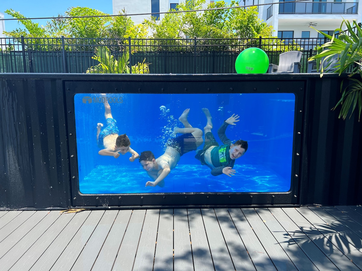 An innovative shipping container pool features clear, blue water, with three individuals swimming inside. Sunlight reflects off the surface of the water, creating a bright, inviting atmosphere. Surrounding greenery and a vibrant green ball enhance the lively outdoor setting.