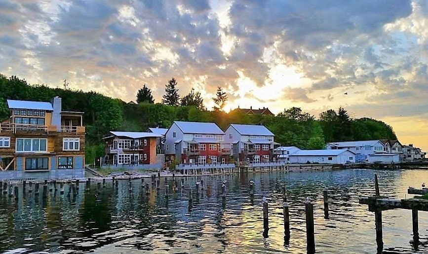 A serene waterfront scene is depicted, featuring a collection of buildings along the shore at sunset. The calm water reflects the colorful sky and structures, while a row of weathered pilings is visible in the foreground, adding to the tranquil atmosphere.
