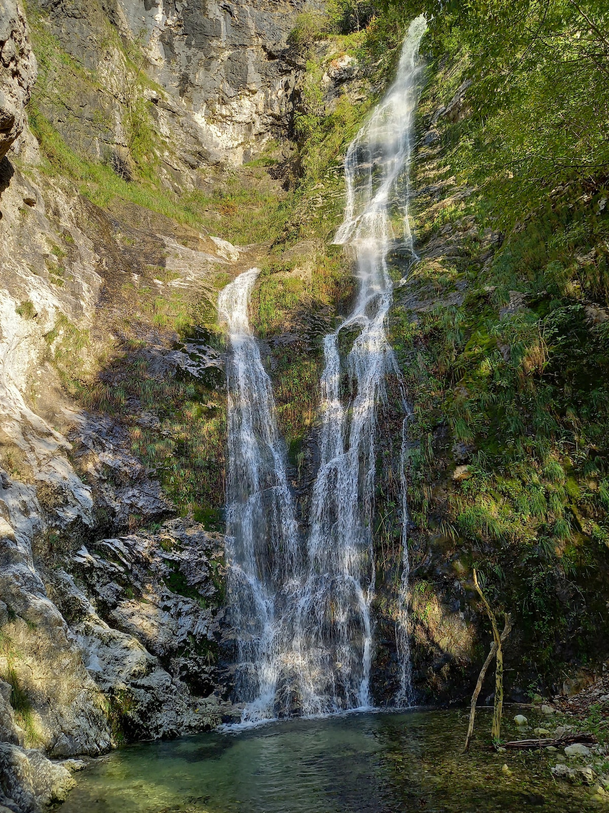 A cascading waterfall is captured amidst rocky cliffs and lush greenery. Water flows down in two prominent streams, creating a serene pool at the base. Sunlight filters through the trees, illuminating the vibrant moss and stones surrounding the water.