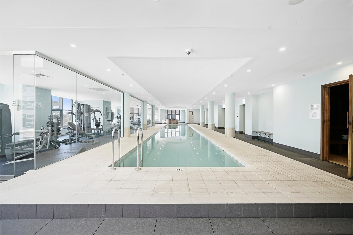 A spacious indoor pool area is complemented by a gym, featuring various exercise equipment. Large glass walls allow natural light to fill the space. The pool's tiled border leads to the calm water, inviting relaxation and fitness opportunities.