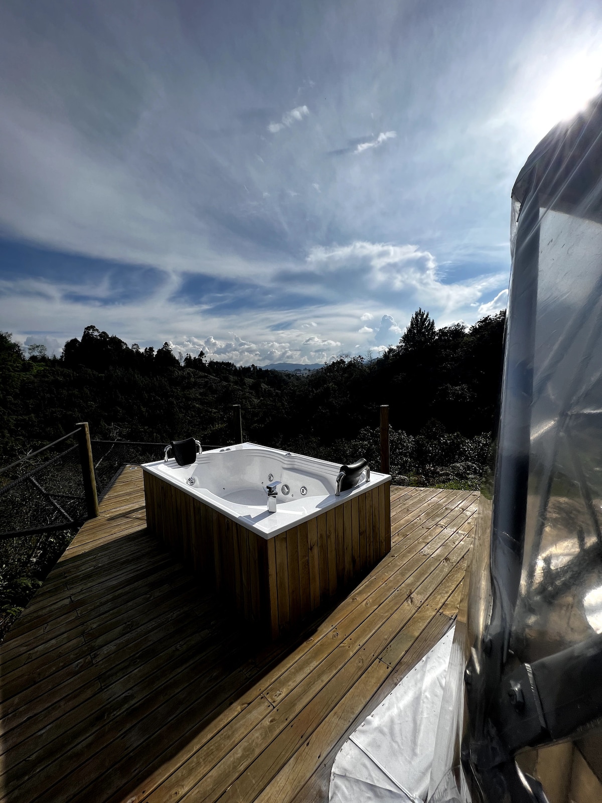 An outdoor jacuzzi with hydromassage features is positioned on a wooden deck, surrounded by lush greenery. The scenic view includes rolling hills and a partly cloudy sky, providing a serene backdrop for relaxation.