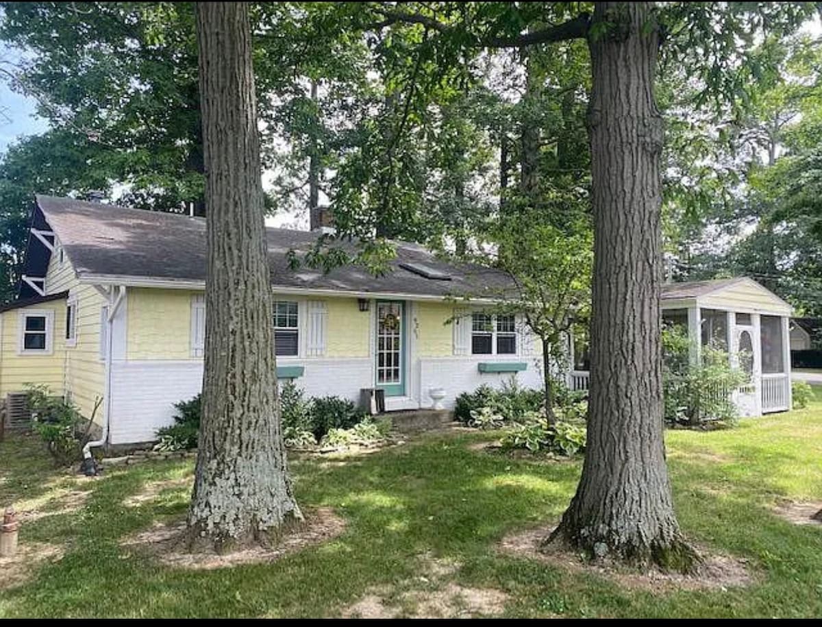 The exterior of the house is framed by two large trees, offering a shaded and serene environment. A light yellow facade is visible, along with bright accents around the windows and entrance. A screened-in patio is located to the right, inviting outdoor relaxation.