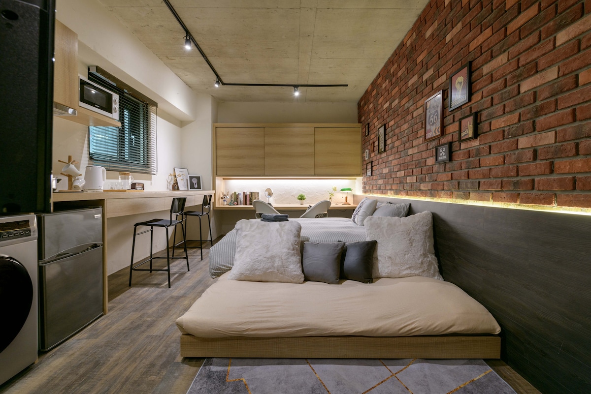 A spacious living area features a comfortable sleeping arrangement with floor cushions and decorative pillows, complemented by warm lighting that outlines the walls. A modern kitchenette is equipped with essential appliances, while a small dining area with a high chair is visible in the background.