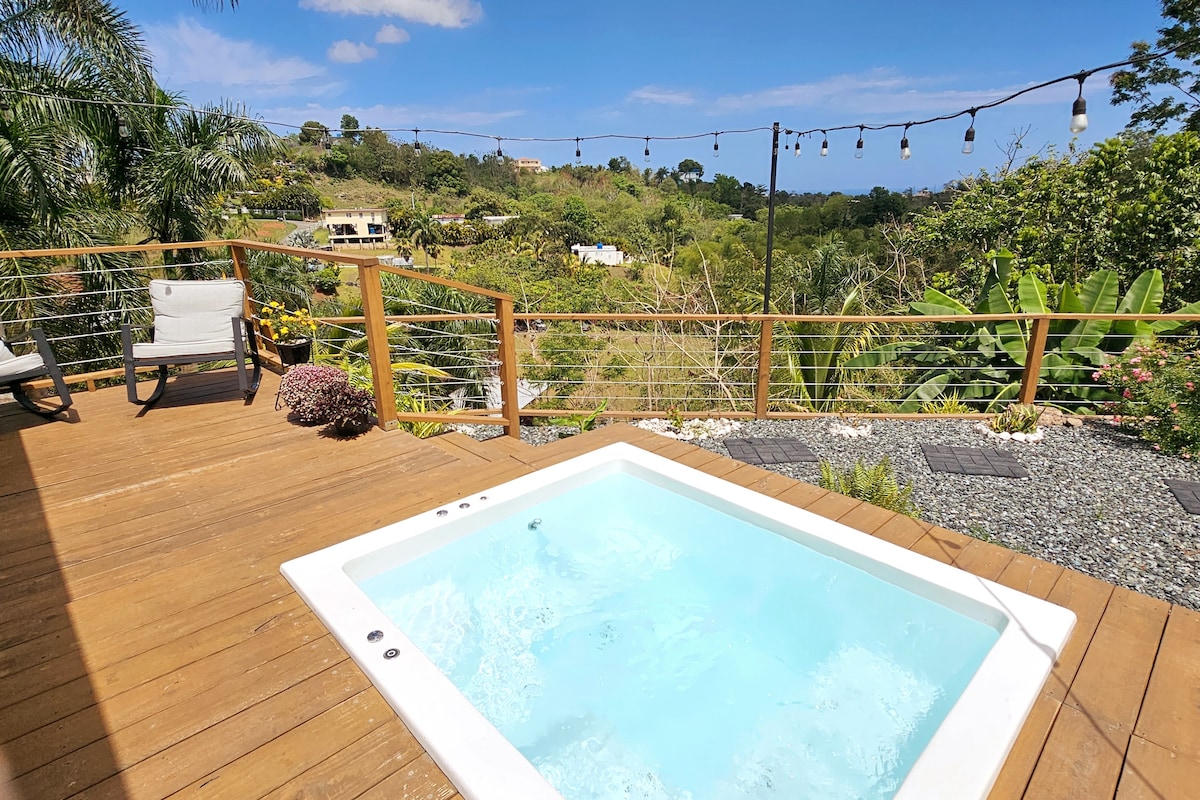 A private outdoor space features a hot tub surrounded by a wooden deck. Lush greenery and a scenic view can be seen in the background, with decorative lights strung overhead. Natural elements enhance the tranquil ambiance, inviting relaxation in a peaceful setting.