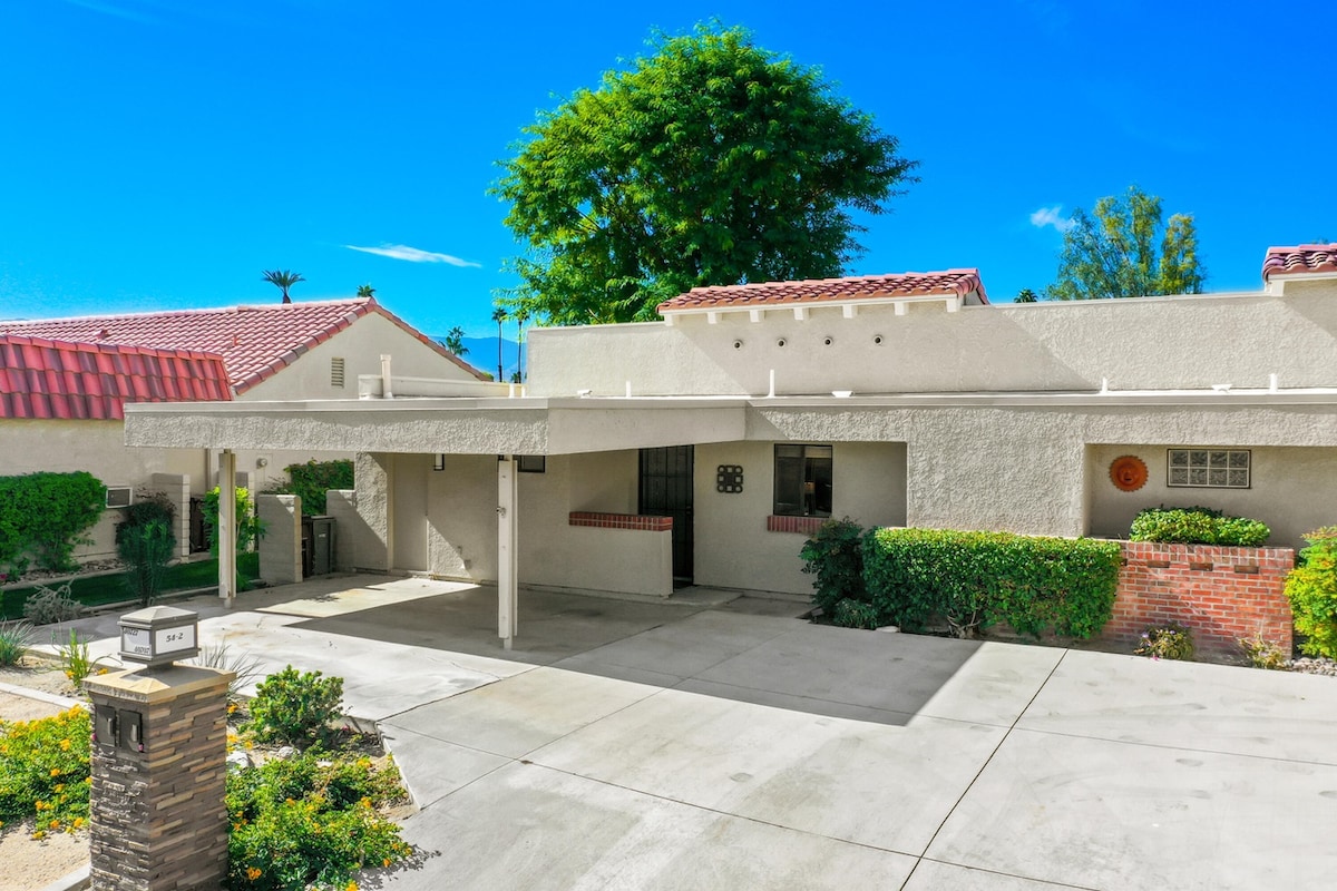 The exterior of the condo is viewed, showcasing a single-level unit with a covered carport. Lush landscaping with green shrubs and trees frames the entrance, while a clear blue sky enhances the inviting feel of the space.