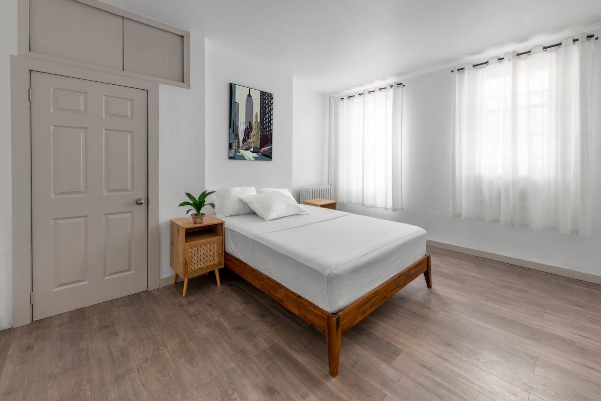A bright and airy bedroom features a spacious bed surrounded by light-colored walls. Natural light filters through sheer curtains, highlighting the wooden bed frame and nightstand. A green plant adds a touch of nature, while a painting adorns the wall, contributing to a calming ambiance.