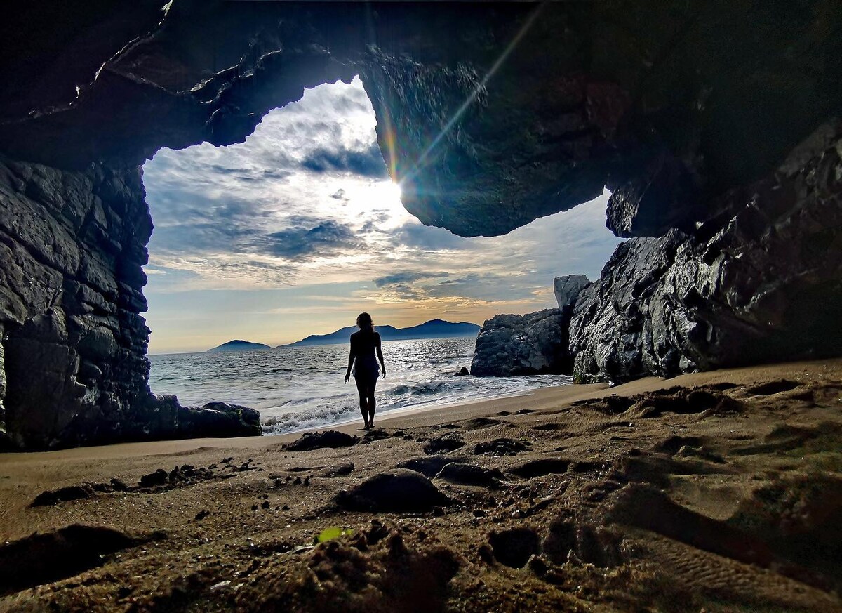A woman walks along a sandy beach framed by large, dark rock formations. The ocean waves gently lap at the shore, and distant islands can be seen against a backdrop of a bright sky with scattered clouds.
