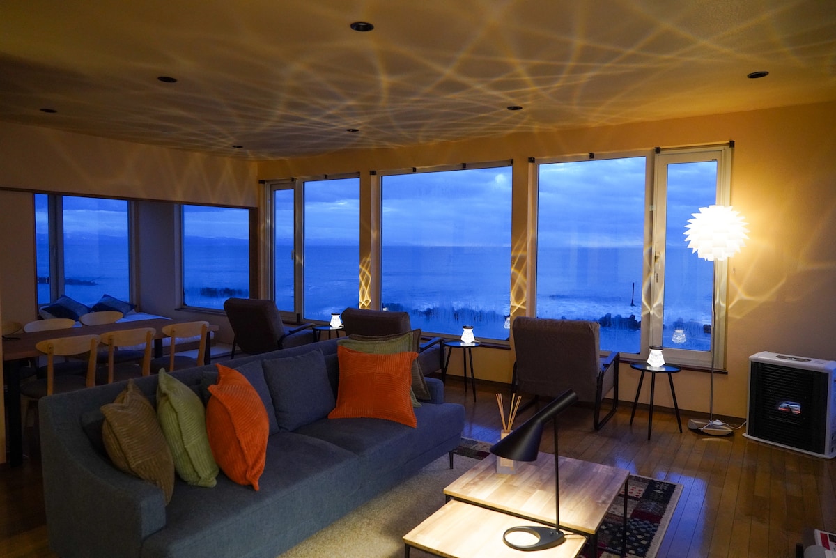 A spacious living area features large windows providing panoramic ocean views as evening light casts soft patterns across the walls. Cozy seating with a mix of cushions is arranged around a modern coffee table, creating a welcoming space for relaxation.