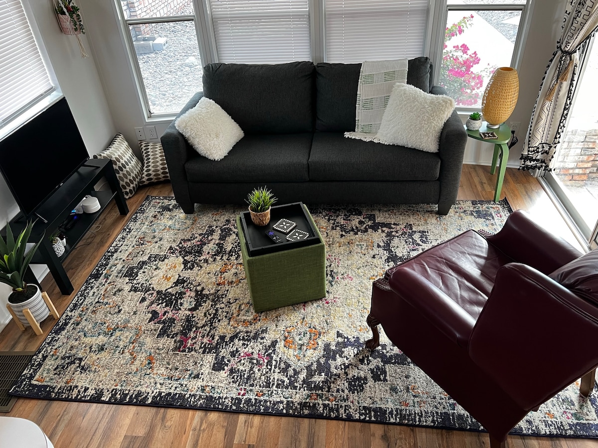 A cozy living area is furnished with a dark sofa and a burgundy armchair, complemented by a colorful area rug. A small green coffee table sits in the center, with a succulent plant on top. Natural light filters through window treatments, enhancing the relaxed atmosphere.