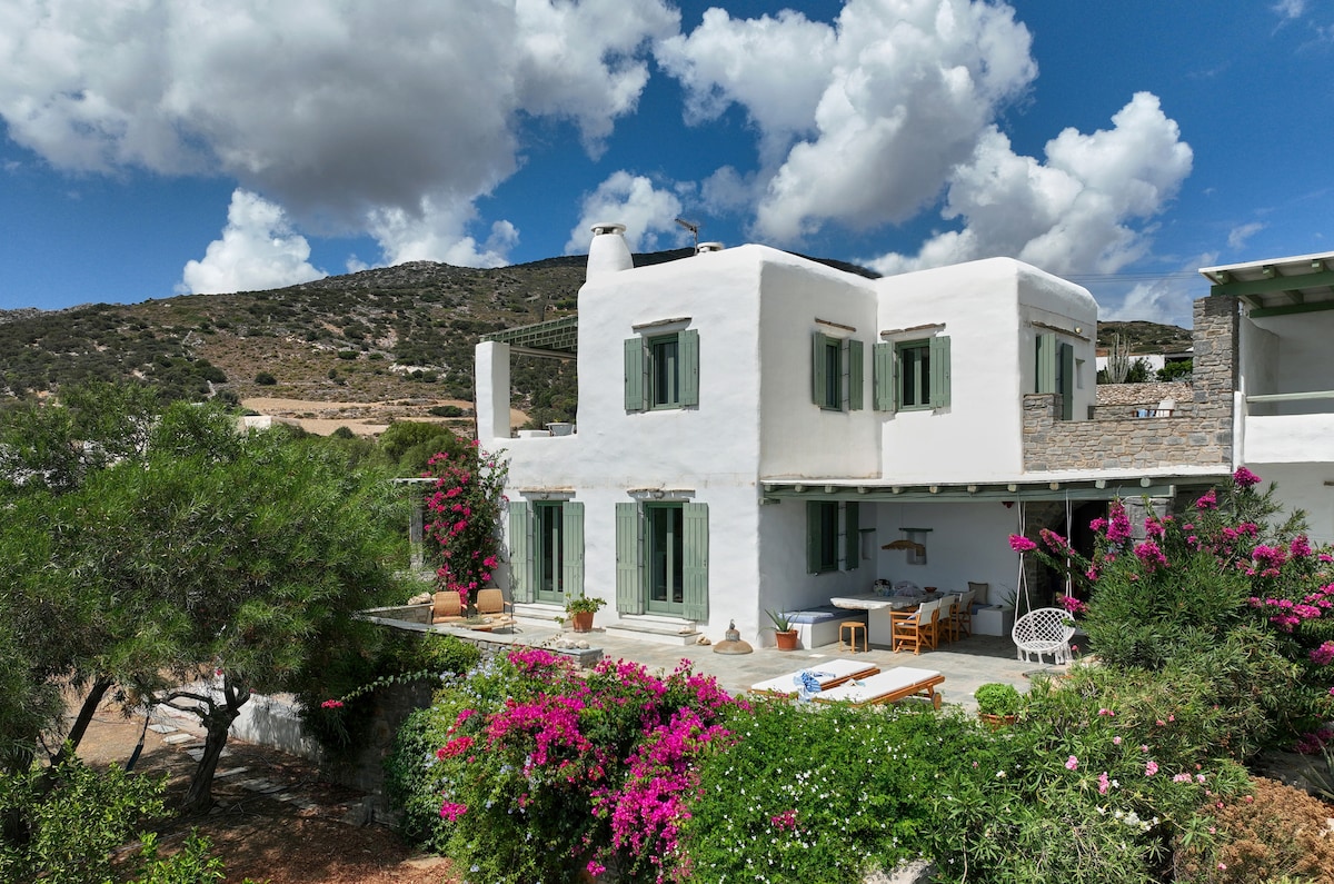 A traditional white house is surrounded by vibrant bougainvillea. Multiple green-shuttered windows provide views of the scenic hillside. An outdoor dining area is visible, inviting gatherings in a serene garden setting.