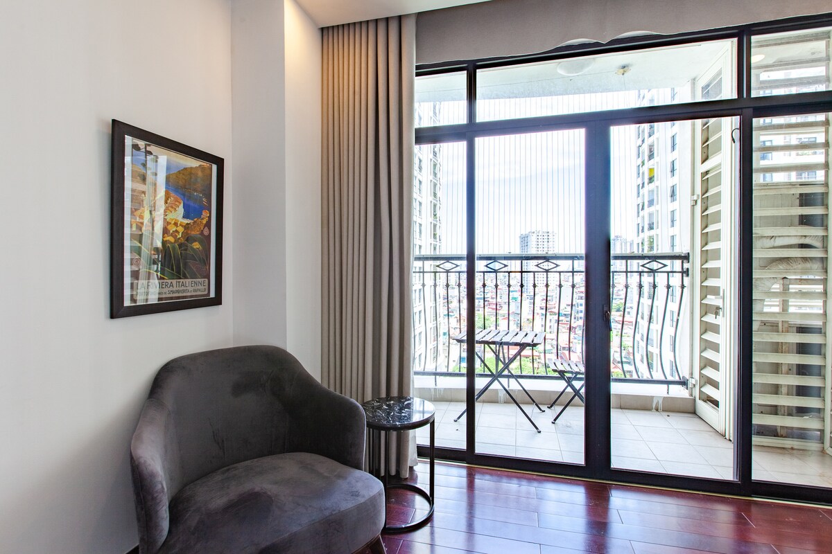 A cozy seating area features a plush armchair beside a small round table. Large glass doors offer a view of the balcony, allowing natural light to fill the space. Floor-to-ceiling curtains frame the entrance to the outdoor area, enhancing the room's openness.