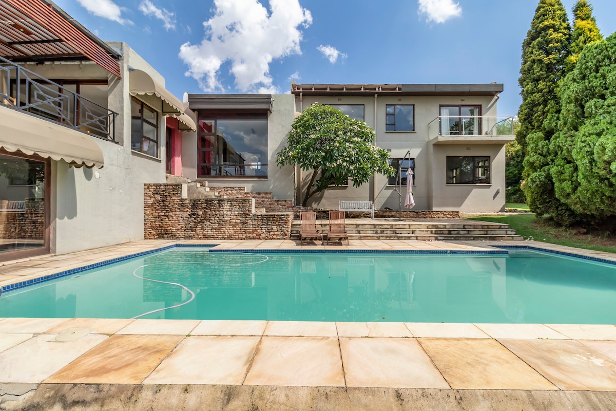 A spacious outdoor area features a clean swimming pool bordered by patio stones. The house stands in the background, showcasing multiple balconies and a shaded tree. Lush greenery surrounds the property, creating a serene environment.