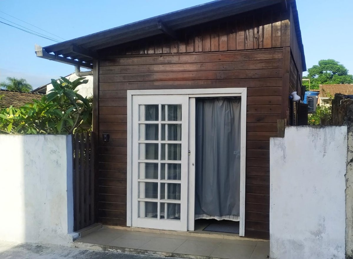 A compact wooden house is presented, featuring a light-colored front door framed by glass panels. A simple curtain hangs inside the doorway, and the exterior walls are textured with natural wood, complemented by a paved stone entrance. Lush greenery is visible beyond the fence.