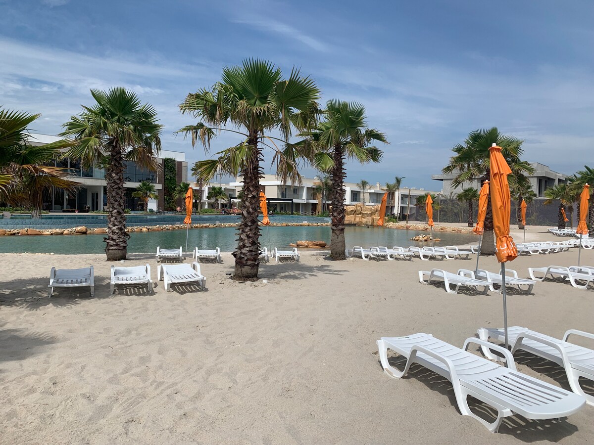 A sandy beach area features several white lounge chairs arranged under palm trees. Bright orange umbrellas add color to the scene, providing shade for relaxation near a tranquil lagoon. The background presents modern buildings, enhancing the contemporary atmosphere.